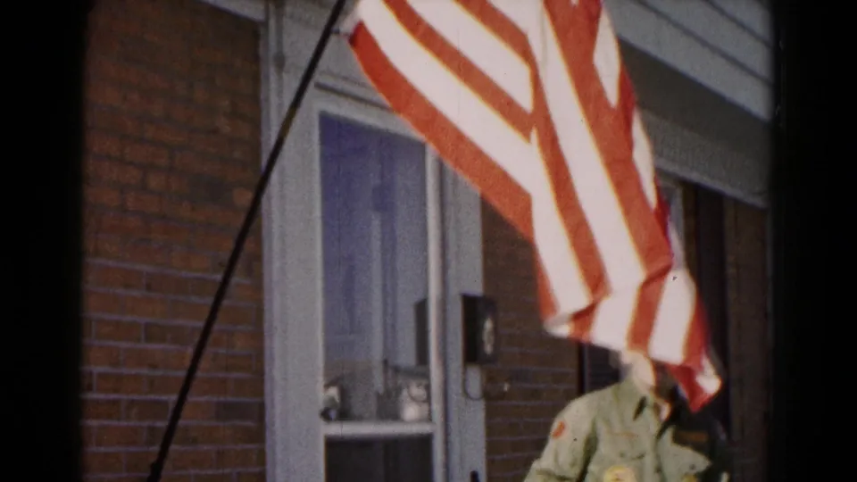 American flag waves close to the camera in front of a brick-and-sided house, with a partially obscured person in green briefly visible near the doorway.