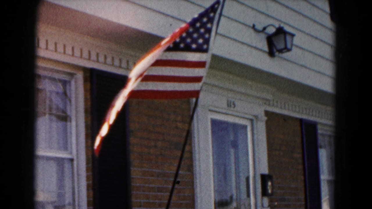 American flag extends from an angled pole outside a residential facade with brick, white siding, black shutters, a porch light, and doorway number 115.