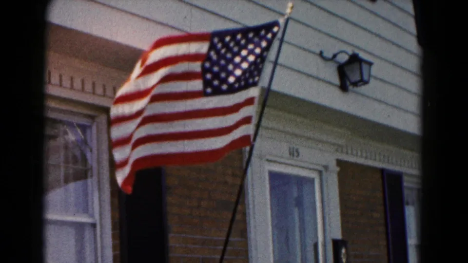 American flag flaps across the foreground beside a brick-and-sided house front, with a window, black shutter, and doorway number 115 visible.