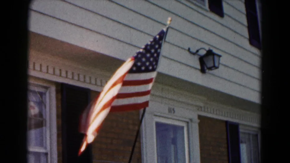 American flag hangs from an angled pole in front of a residential exterior with white siding, brickwork, a black shutter, a porch light, and the number 115 near the door.