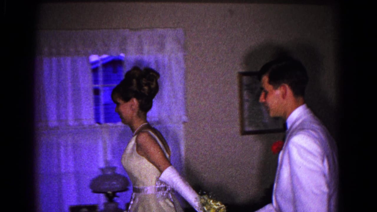 Young woman in formal dress and gloves walks ahead of a young man in a white tuxedo indoors.