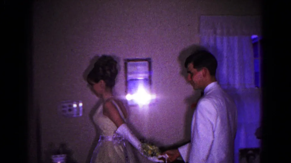 The young woman in gloves and a formal dress walks past wall decor while the young man in a white tuxedo follows behind her.