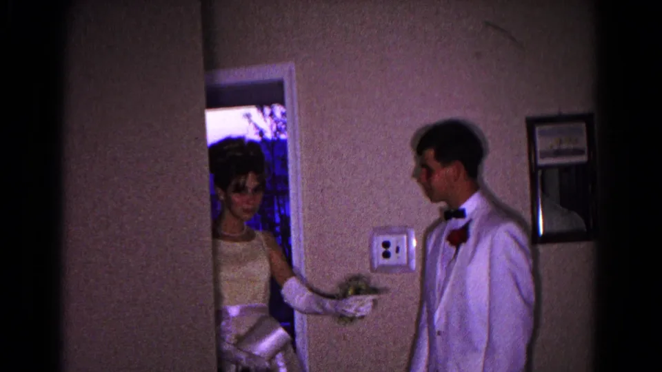 A prom couple pauses by a doorway, with the young woman holding flowers and the young man in a white tuxedo looking toward her.