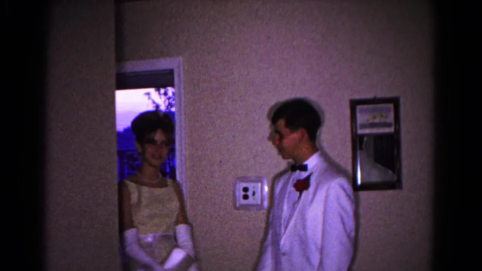 The prom couple shares a smile in a private interior, with evening light visible through the doorway behind the young woman.