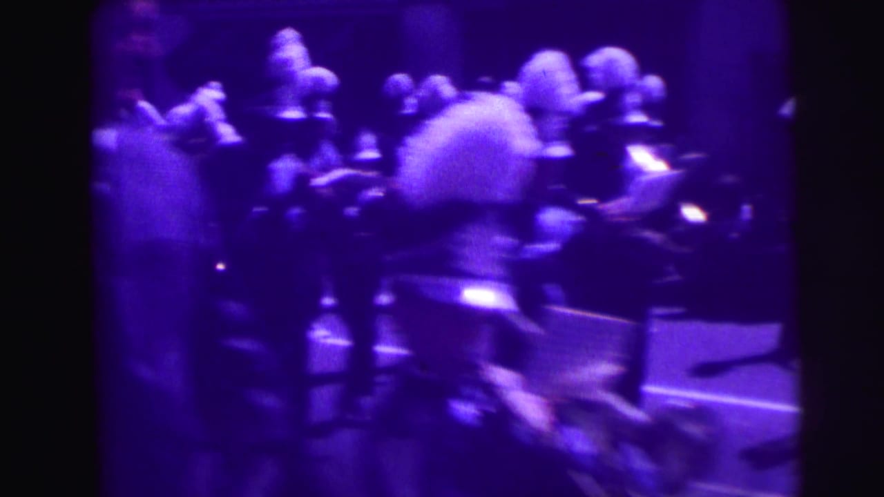 Uniformed marching band members moving along a street in purple-tinted 8mm footage.