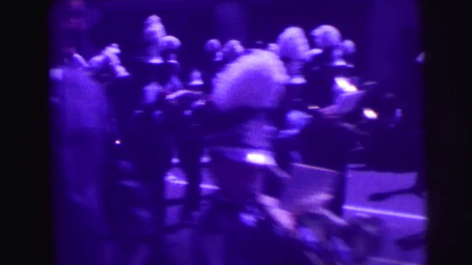 Purple-tinted 8mm frame of uniformed band members and musicians passing through a street parade, identified by metadata as 1969.