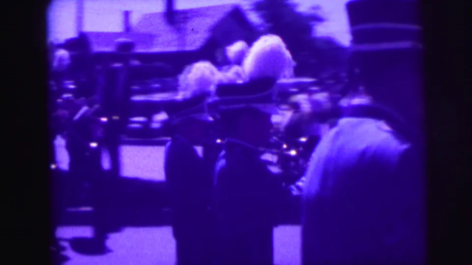 A purple-tinted 8mm frame shows a marching band member near a drum and other parade participants on the street.