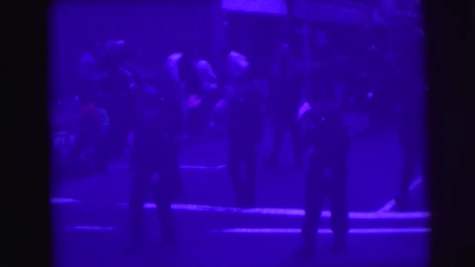 Purple-tinted 8mm frame showing adults or officials standing on a parade route with a group of participants in the background.