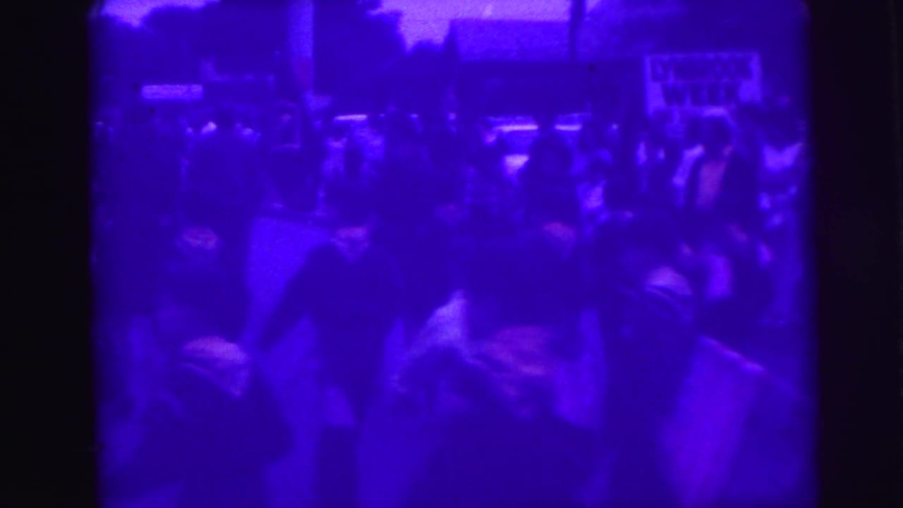 1969 home-movie frame from Lynbrook metadata showing spectators watching a street parade with American flags visible in the crowd.