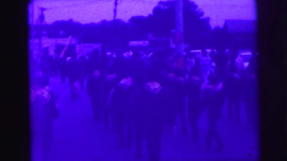 Home-movie frame showing a small-town parade scene with American flags, roadside viewers, and soft 8mm grain.