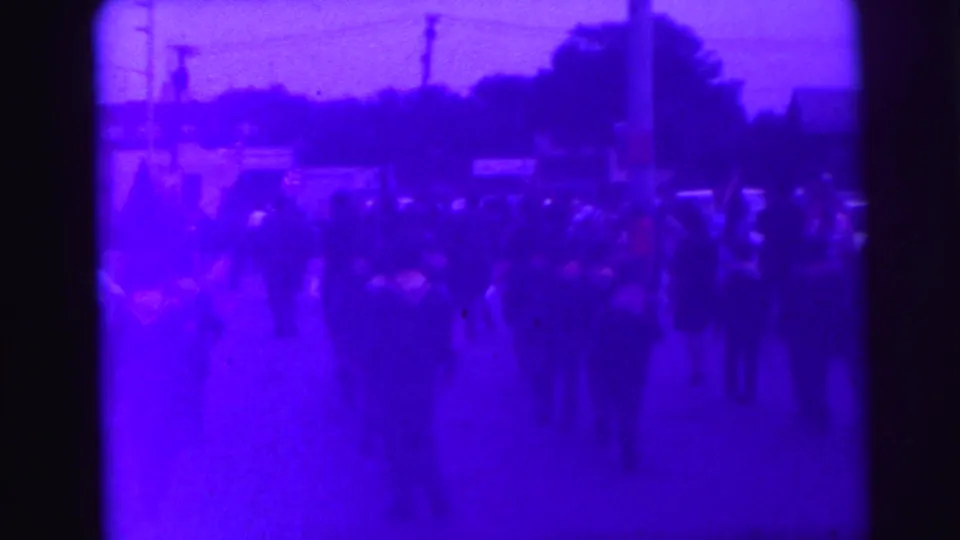 Late-1960s Lynbrook parade frame, based on supplied metadata, with flags and spectators visible in a soft purple 8mm image.