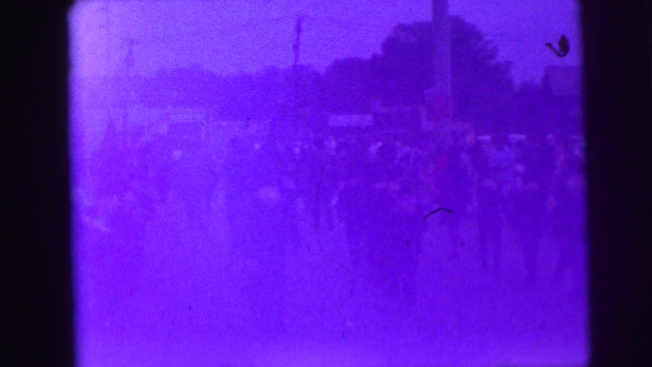 Soft, purple-tinted 8mm frame showing a distant group of people in an outdoor public setting, with heavy blur and film wear.