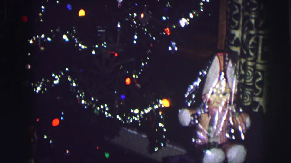 Handheld 8mm frame showing a lit Christmas tree, shiny tinsel, ornaments, and presents gathered near the base.