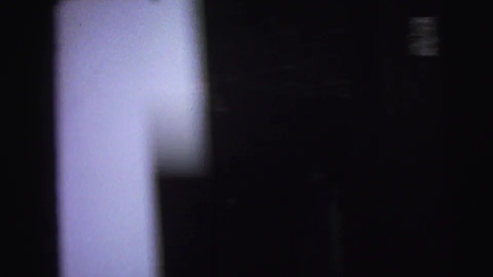 Underexposed 8mm frame of a dark private interior, with a pale blurred vertical shape at left and little detail elsewhere.