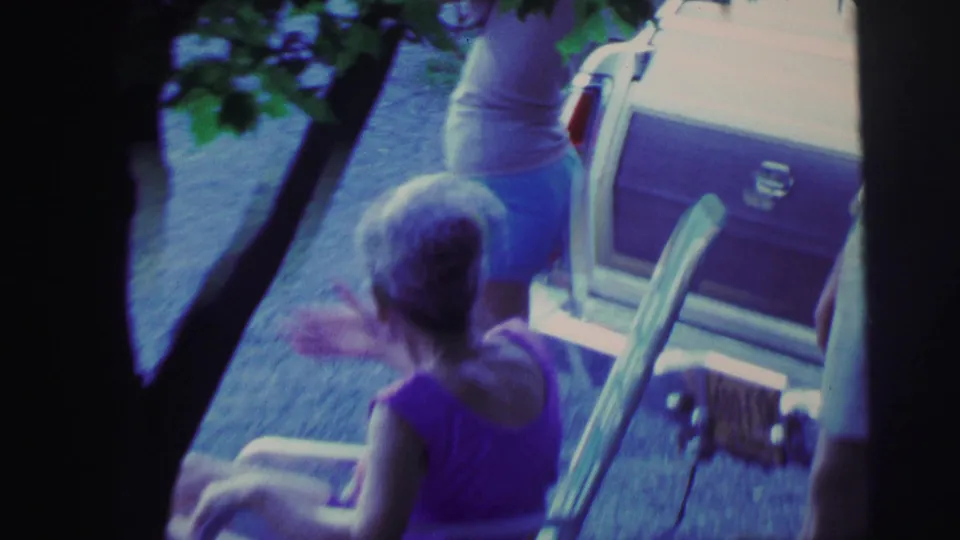 1971 8mm home-movie frame of an older woman seated in a folding chair while adults stand beside a parked station wagon under leafy shade.