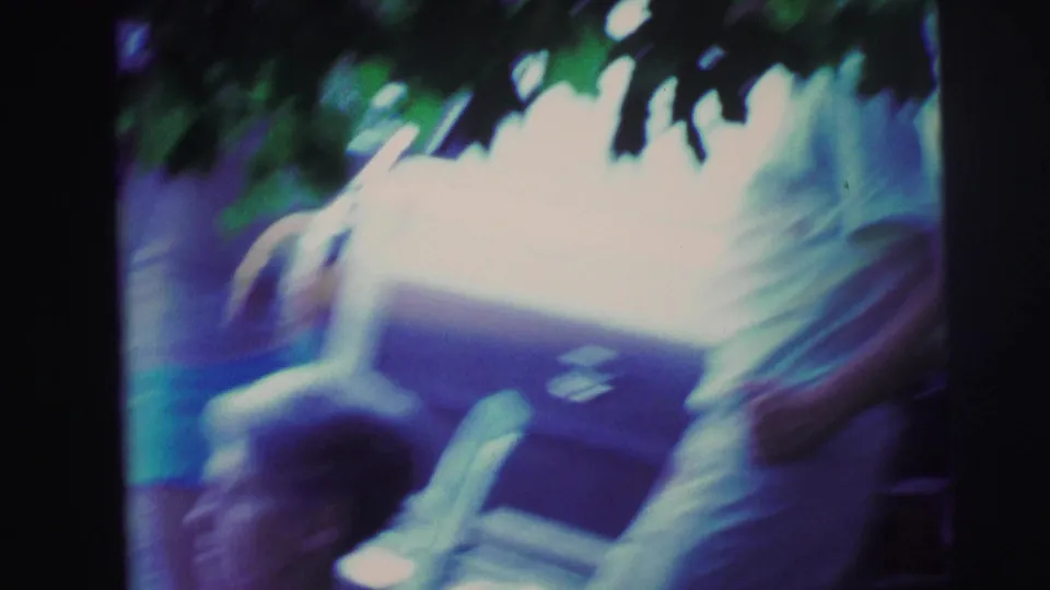 Soft, motion-blurred 8mm frame of adults standing near a parked station wagon, with the seated older woman partly visible at the bottom edge.