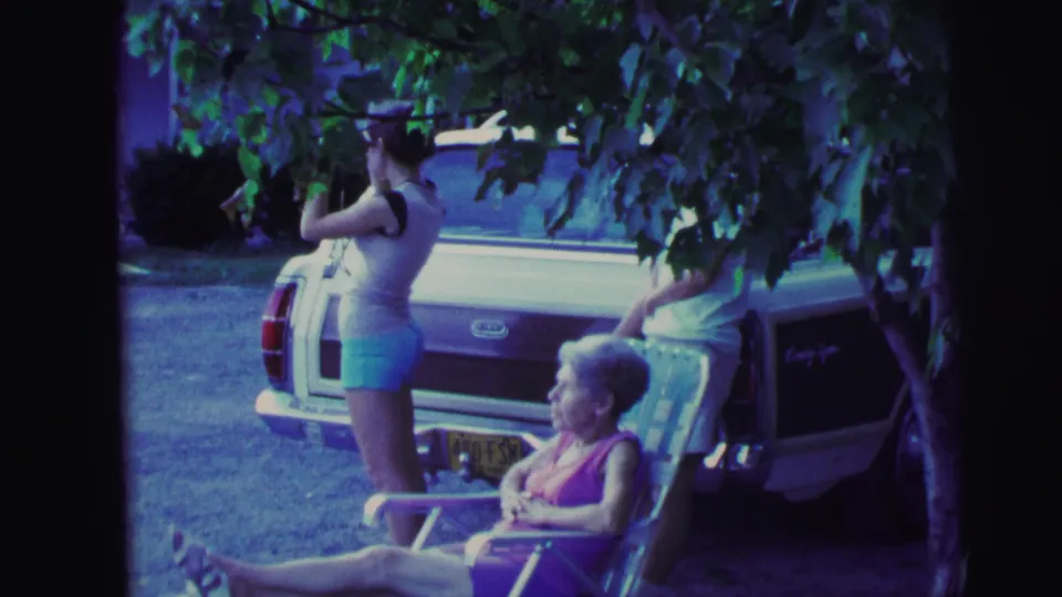 Wider 1971 8mm frame of an older woman in a folding chair and two adults standing at the rear of a white and maroon station wagon under trees.