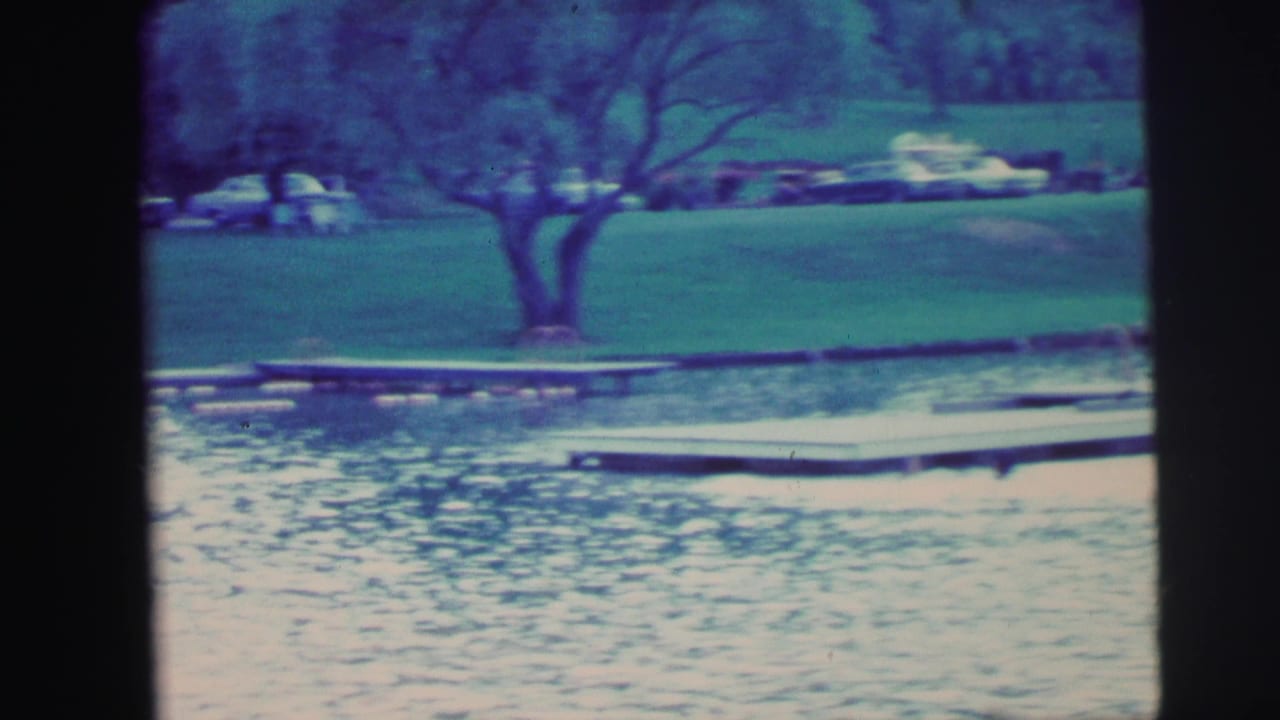 1971 8mm home-movie frame showing a lakeside dock, lawn, trees, and several parked cars across the water.