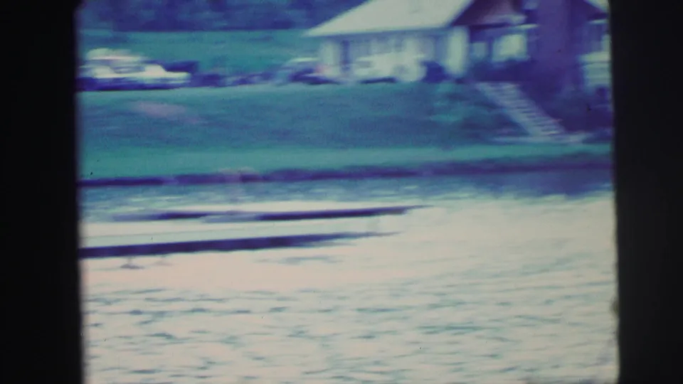 Soft 8mm frame of a dock along the water with a house, lawn, trees, and parked cars visible across the lake.