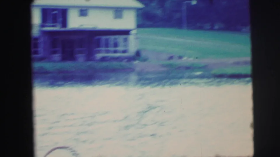 1971 8mm frame showing a lakeside house, dock, open lawn, and small indistinct figures or objects near the waterline.