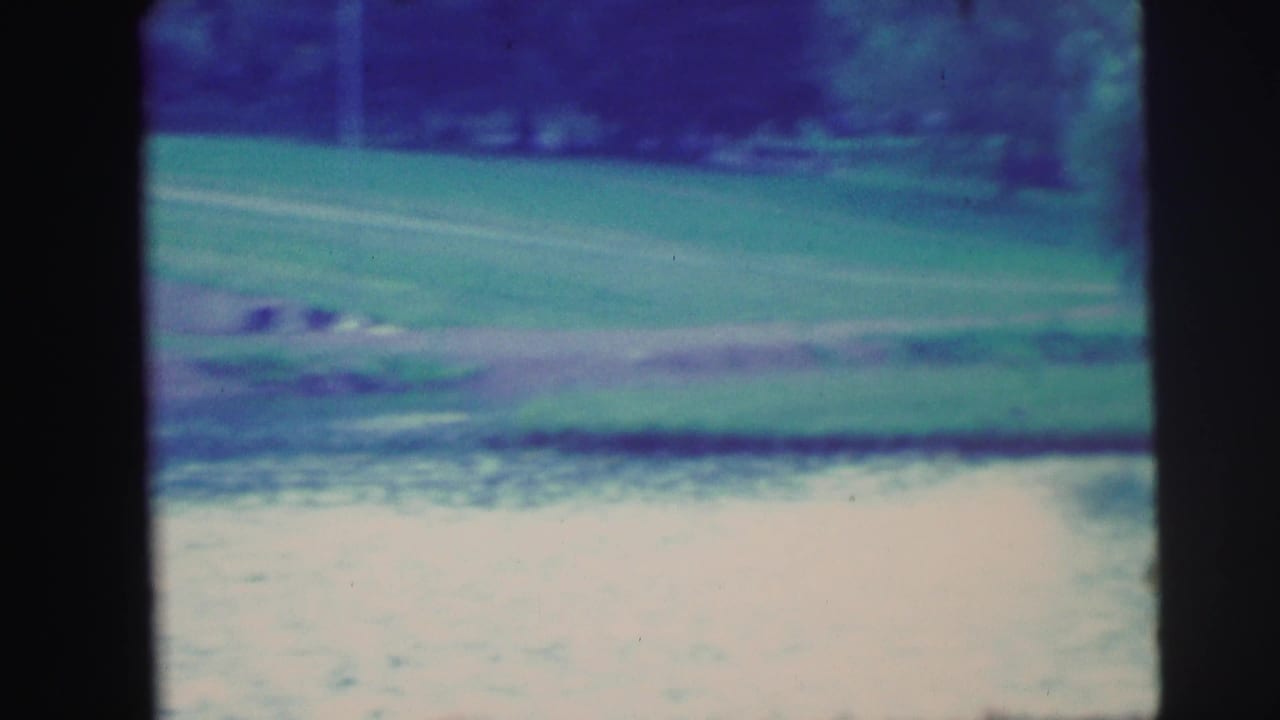 1971 8mm frame showing a quiet grassy lakeshore, pale water, trees, and a distant bank in soft blue-green color.