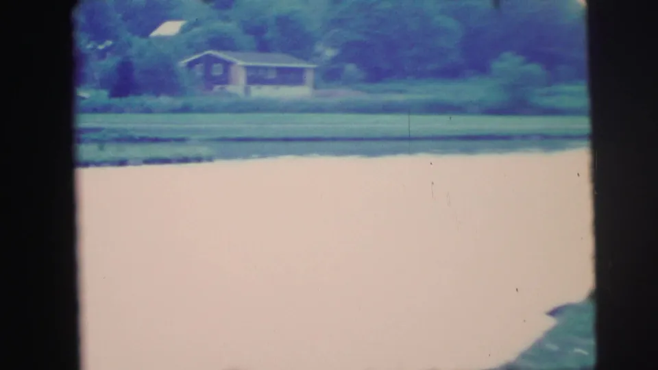 1971 8mm frame looking across still water toward a small house, lawn, and tree line on the opposite shore.