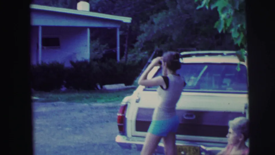 1971 8mm frame showing a woman in shorts and a tank top using binoculars beside a parked station wagon near a house and trees.