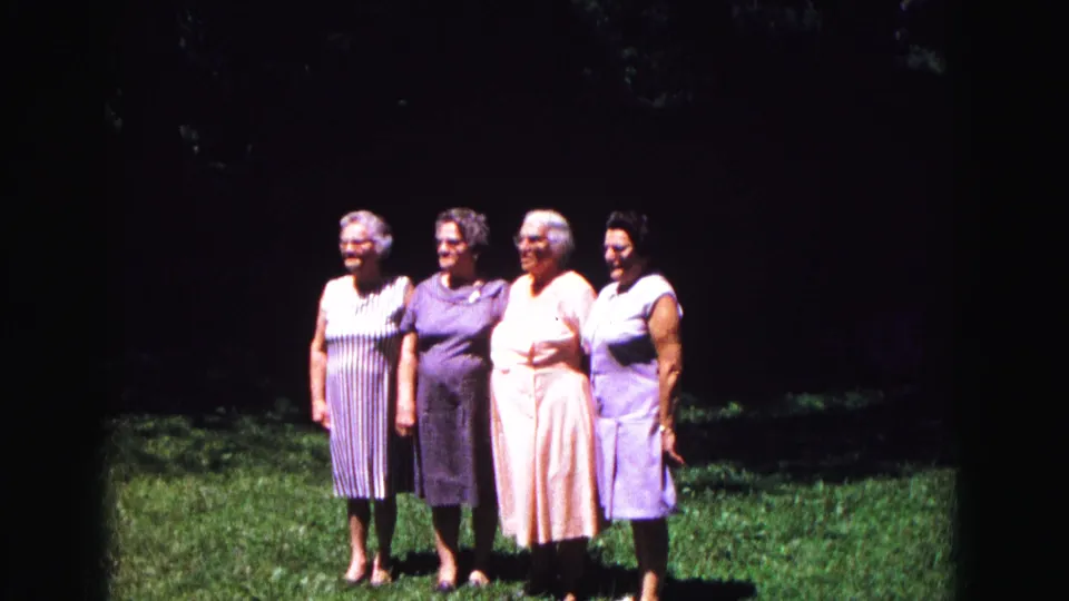 Four older women in striped, purple, peach, and light dresses pose together outdoors in 1971 home-movie footage.