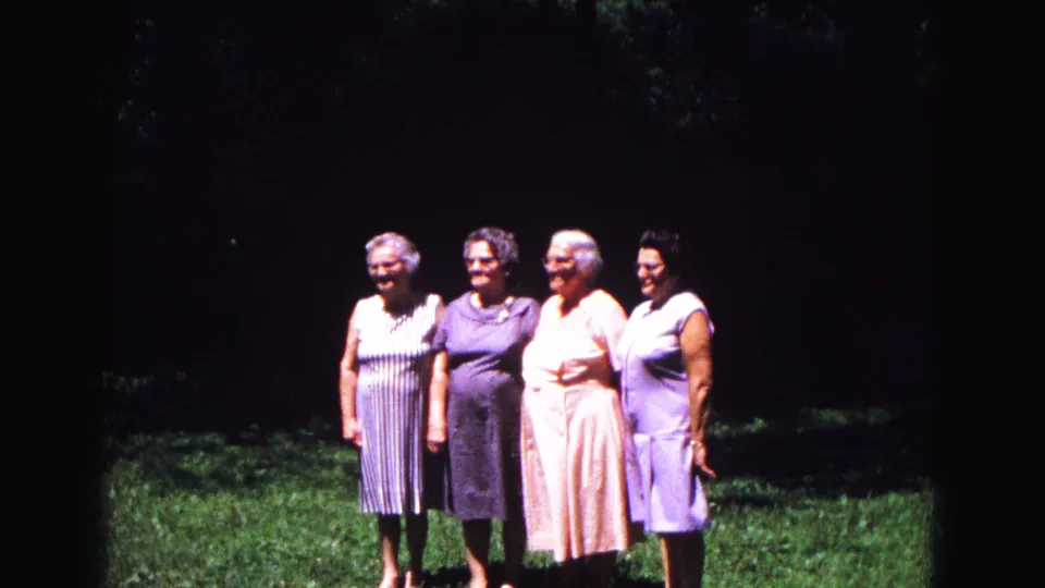 Four older women stand shoulder to shoulder in an outdoor home-movie shot, with colorful dresses and a dark wooded background.