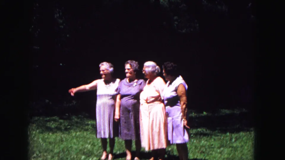 At the end of the clip, the woman at left gestures off-camera while the group turns slightly, still gathered on the grass.