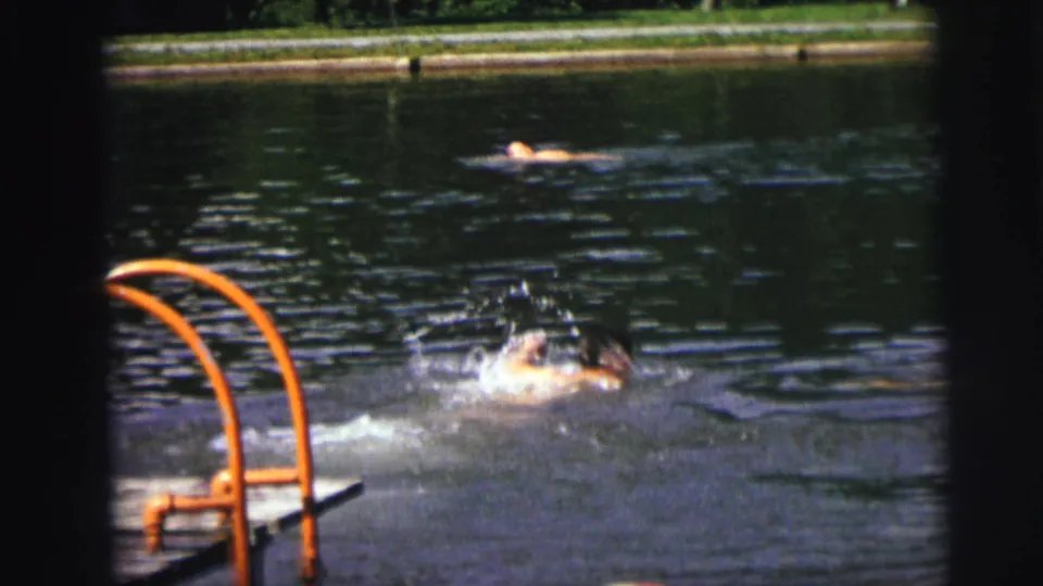 1972 8mm frame with a swimmer surfacing in dark water near an orange ladder and another swimmer in the distance.