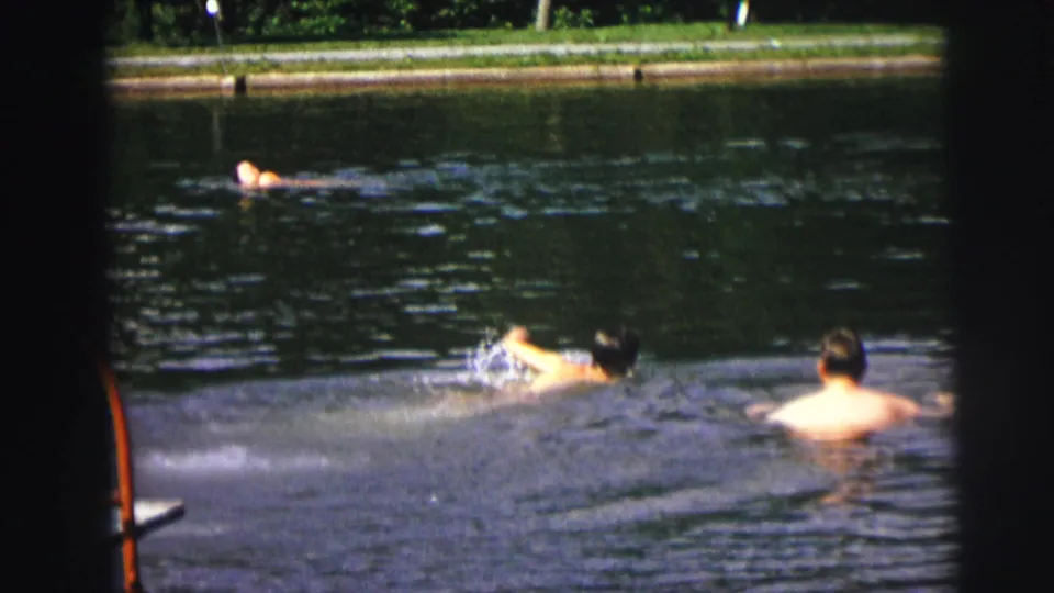 1972 8mm frame showing one swimmer crossing the foreground while another bare-backed person watches from the water.