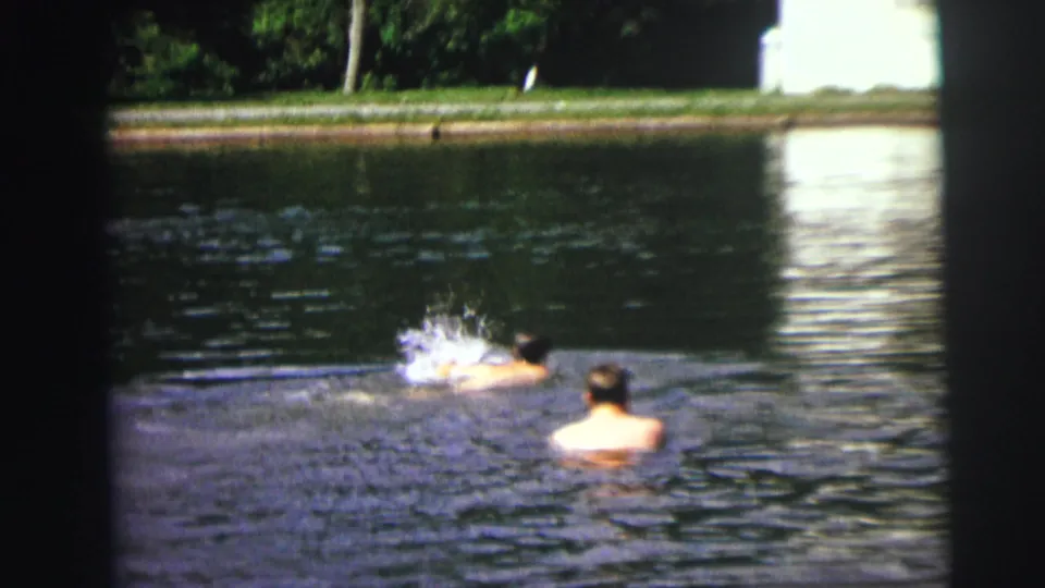 1972 8mm frame of a swimmer creating splashes while another person faces them from the pool water.