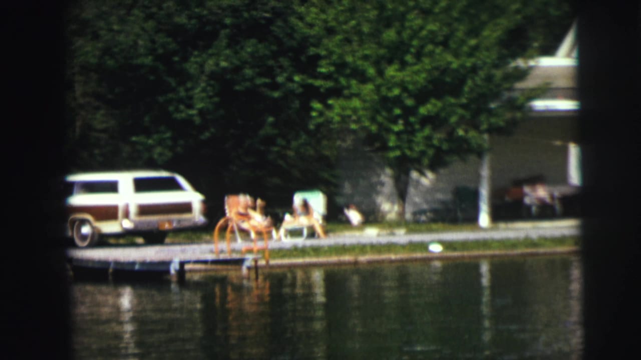 Station wagon parked beside a dock with lawn chairs and people near calm water.