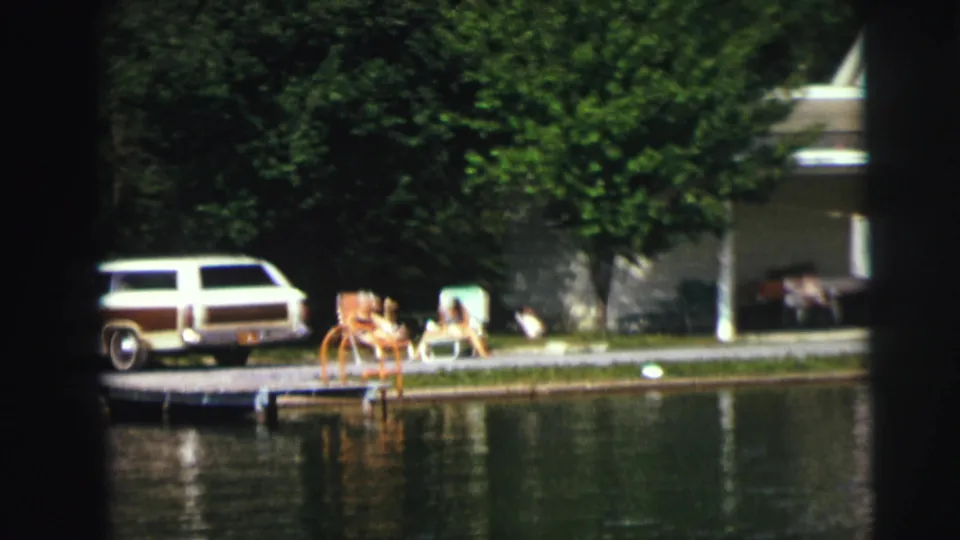 Soft 8mm frame of a wood-paneled station wagon near a small dock, with people seated on grass beside reflective water.