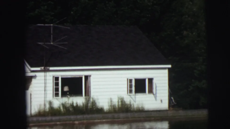 Soft frame of a light-colored waterside house with open windows, a dark roof, trees behind it, and water in the foreground.