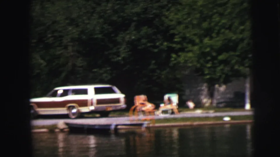 Wide 8mm view of the station wagon and dockside seating area, with people near the chairs and dense trees behind the shoreline.