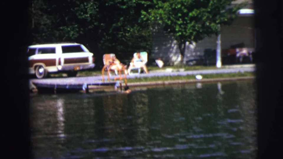 1972 8mm frame showing people relaxing by a lakeside dock as a swimmer is near the water and a wood-paneled station wagon sits behind them.