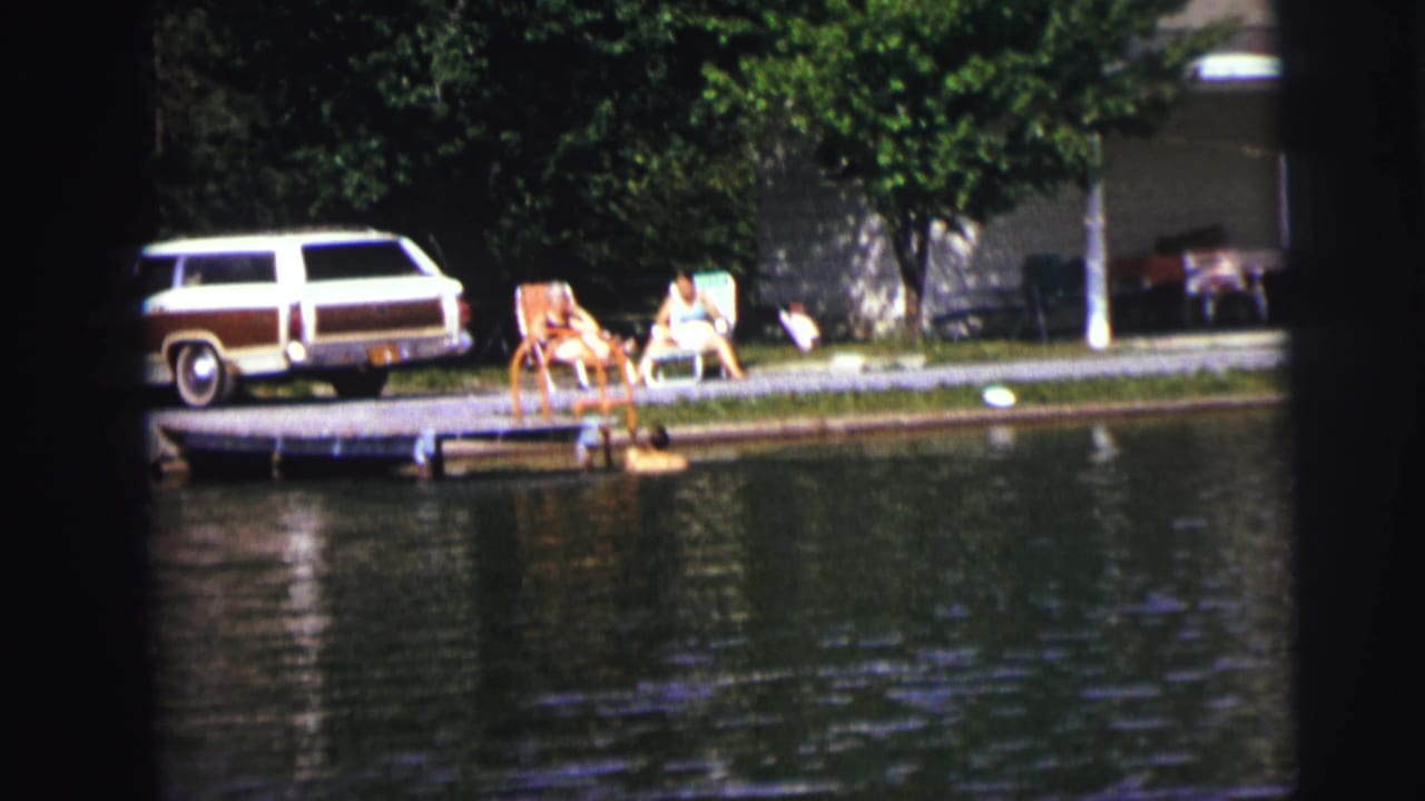 A swimmer near the dock with two seated people and a station wagon behind the shoreline.