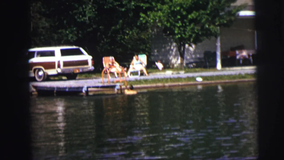 Home-movie frame of a swimmer at a lakeside dock while two people sit in folding chairs near a parked wood-paneled station wagon.