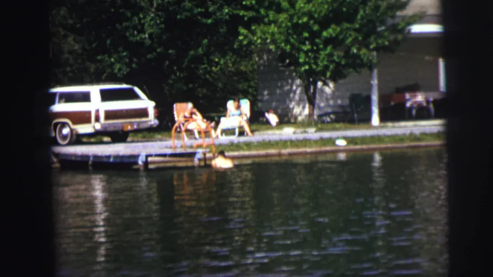 1970s lakeside home-movie frame with a swimmer climbing or resting near the dock as seated people watch from the grassy shore.