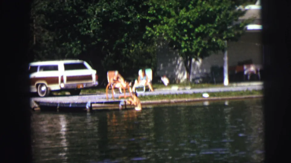 Soft 8mm frame of a person at the lakeside dock while others lounge in chairs and a wood-paneled station wagon remains parked nearby.