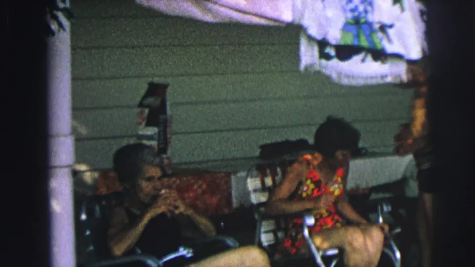 1970s 8mm home-movie frame of two women in swimsuits seated outside a home, with a man standing near white siding.