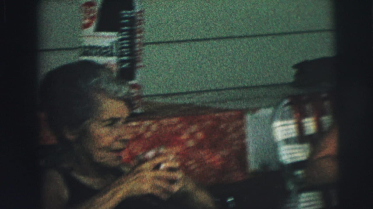 A seated woman drinks while the camera frames her near house siding.