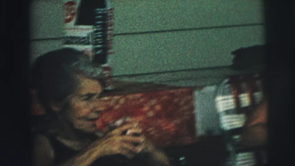 Close, soft 8mm frame of a woman seated outside near white siding, raising a drink or cup toward her mouth.