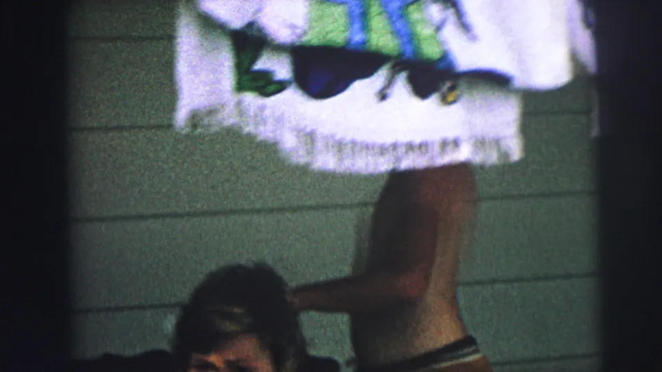 Handheld 8mm frame showing a shirtless man in swim trunks near hanging towels, with part of a seated woman below.