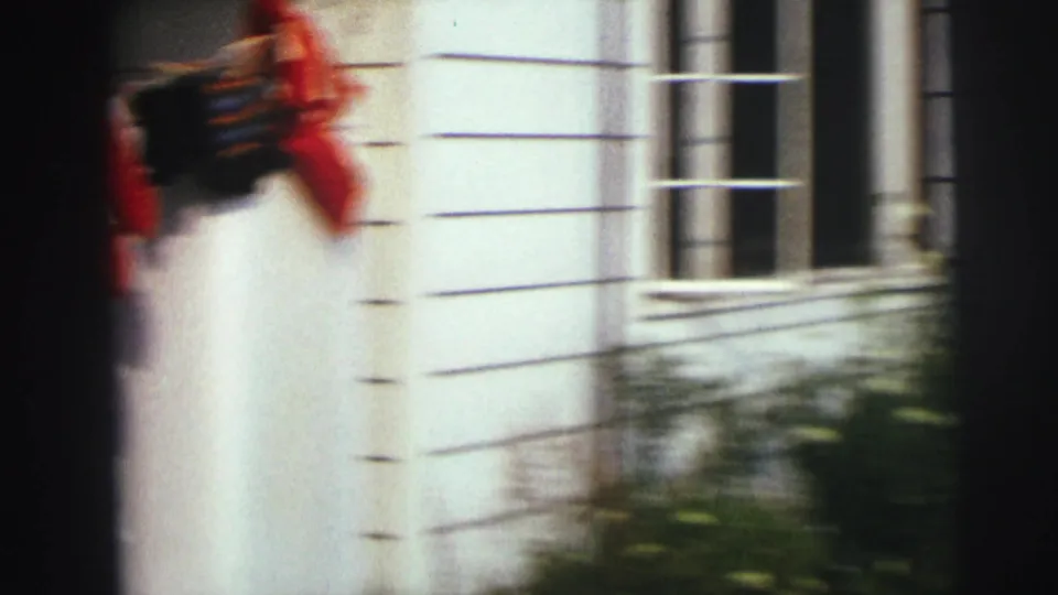 Blurred handheld 8mm frame of a house exterior with white siding, a window, plants, and a red object passing at frame edge.