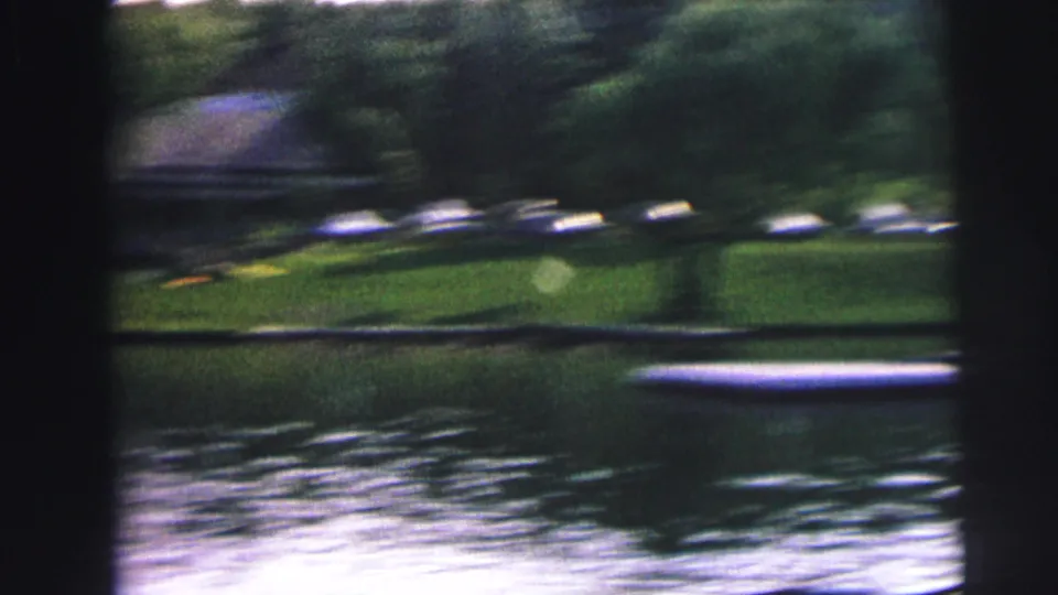 Soft, handheld 8mm view across a lake toward a grassy shore with trees and several cars, stated as 1972 Mobile Alabama context.