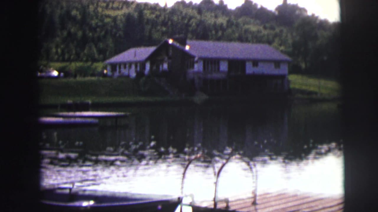 Lakeside house across calm water with dock, boat, trees, and wooded hill.