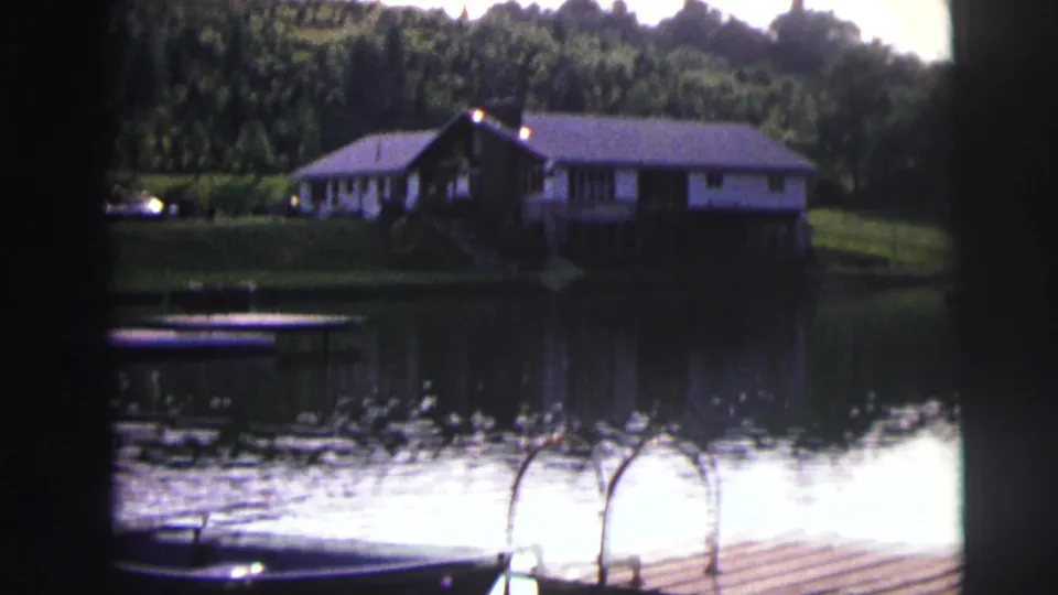 A lakeside house stands beyond calm water, with a dock, small boat, and wooded shoreline in archival 8mm footage.